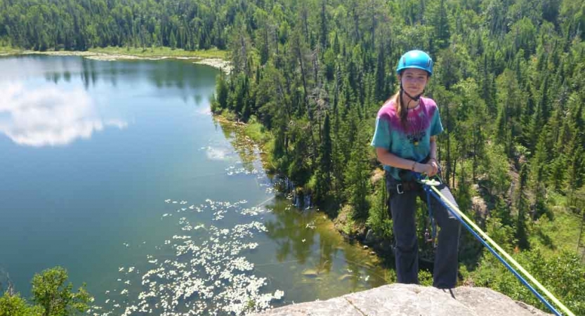 A young person wearing safety gear is secured by ropes as they stand on the edge of a cliff, high above a body of water and a forrest. 